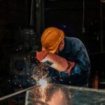 A welder wearing protective gear works on metal in an industrial workshop, surrounded by sparks.